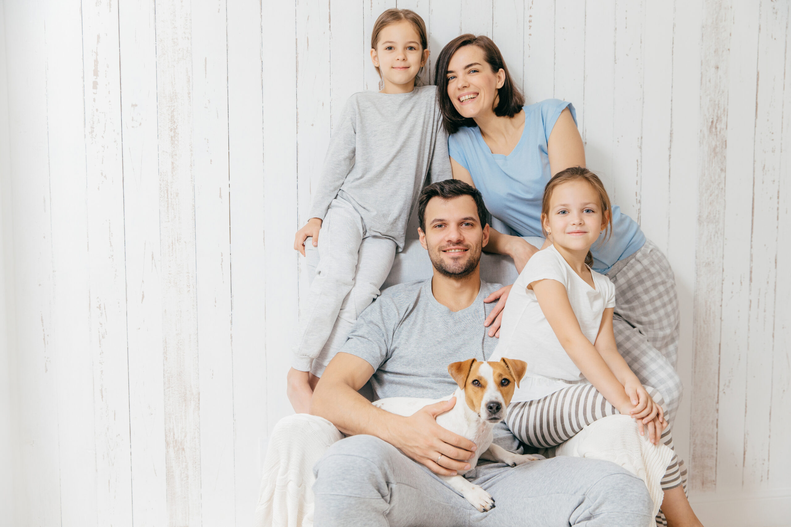 Horizontal shot of friendly family pose together against white background: two little sisters, father, mother and their pet. Happy parents and their female children. Family of four. Parenthood